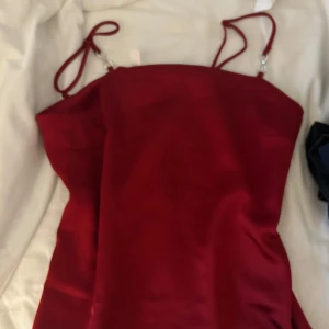 Röd topp med strassdetalj från H&M - Säljer en röd, figurnära topp från H&M med smala axelband och snygga strassdetaljer. Toppen har en enkel, elegant design och är tillverkad i ett glansigt material som ger en lyxig känsla.