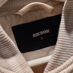 Beige bomberjacka från BikBok - Cremefrägad, lite glansig bomberjacka från BikBok i storlek S. 