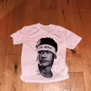 Vit t-shirt med grafiskt tryck och text - Vit t-shirt med stort svart tryck av ett porträtt på framsidan, där personen har ett pannband med texten '100% JESUS'. Baksidan har ett stort svart sköldmönster med pilar och stjärnor. Rund halsringning och korta ärmar.