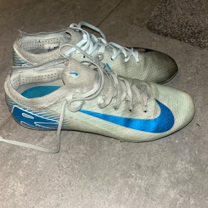 Nike fotbollsskor med blå detaljer - Säljer ett par Nike fotbollsskor i vitt med blå swoosh och detaljer. Dom är äkta och köpt på unisport. Ända skadan är att det lossnat en dobb på varsin sko. Men det går att spela med ändå, har själv använt dom i match och träning i detta skick. Kan skicka på kvittot från unisport om det önskas.