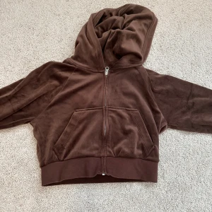 Brun hoodie i sammet - Mysig brun hoodie i mjuk sammet med dragkedja framtill och stor huva. Tröjan har två fickor på magen och bred mudd nedtill. Perfekt för en avslappnad stil.
