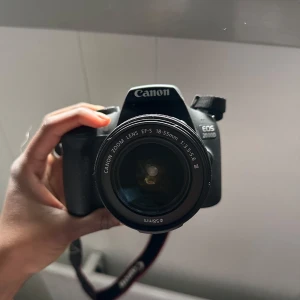 Canon EOS 2000D systemkamera med 18-55mm objektiv - Säljer en svart Canon EOS 2000D systemkamera tillsammans med ett Canon EFS 18-55mm objektiv. Kameran har tydliga knappar och display på baksidan samt en klassisk kamerarem. Perfekt för dig som vill börja fotografera med systemkamera. Du får med batteri laddaren och ett tomt missekort !!