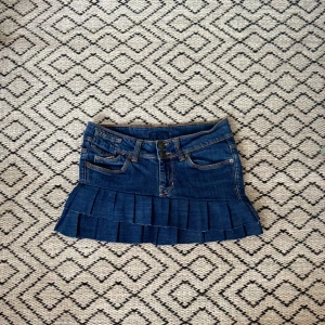 Lågmidjad jeanskjol med volanger - Blå jeanskjol med volang nertill. Kjolen har klassiska fickor fram och bak, dubbla knappar och synliga sömmar. Perfekt för dig som gillar en trendig stil. Omsydda jeans från Gina, noggrant! 