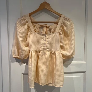 Beige rutig blus med puffärmar och knytning - Supersöt beige blus med rutigt mönster, puffiga trekvartsärmar och knytning i ryggen. Djup v-ringning och knappar framtill ger en romantisk vibe. Perfekt till jeans eller kjol!