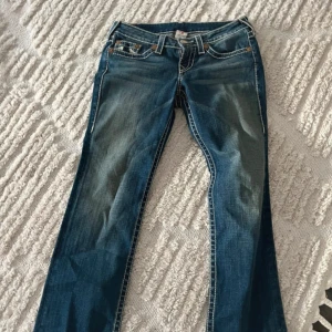 Blå bootcut jeans från True Religion - Säljer ett par klassiska blå jeans från True Religion med bootcut passform. Jeansen har tydliga kontrastsömmar, broderade bakfickor och en ikonisk patch bak i midjan. Perfekta för dig som gillar en tidlös jeanslook.