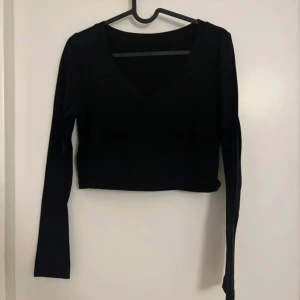 Svart croppad långärmad topp - En svart croppad topp med lång ärm och v-ringning. Enkel och stilren design som passar till många olika outfits. Perfekt för dig som gillar en minimalistisk look.