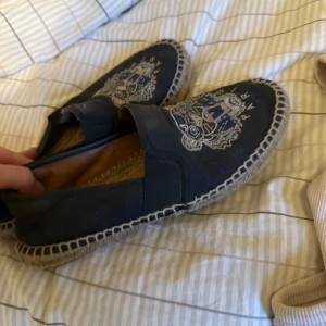 Svarta espadrillos med broderad tiger - Säljer ett par svarta espadrillos från Kenzo med broderad tiger och logga på ovandelen. Skorna har en klassisk flätad sula och är tillverkade i textil. Perfekta för sommaren och ger en cool touch till din outfit.
