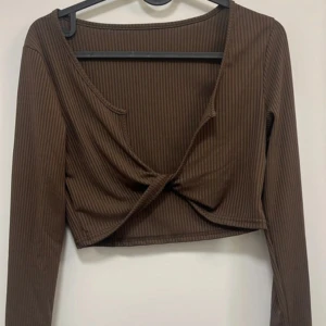 Brun croppad omlottopp - Säljer en brun croppad topp med omlottdesign och lång ärm. Toppen har diskreta ribbade ränder och en twistad detalj framtill. Perfekt att styla med höga jeans eller kjol.