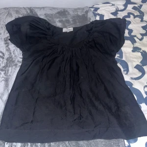 Svart blus med puffärmar - Svart blus med rundad halsringning och korta puffärmar. Lös passform och lätt rynk vid halsen för en luftig känsla. Perfekt för en stilren och enkel look.