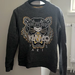 Mörkgrå sweatshirt med tigertryck från Kenzo - Cool mörkgrå sweatshirt från Kenzo med stort tigertryck och broderad logga på bröstet. Tröjan har rund halsringning och långa ärmar. Perfekt för dig som vill sticka ut med en ikonisk design.