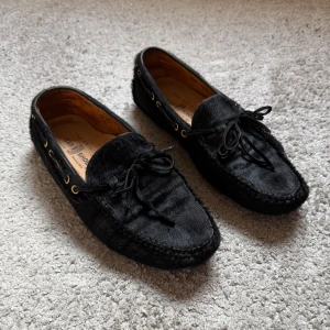 Svarta loafers från Car Shoe - Nypris 7800kr Säljer ett par svarta loafers från Car Shoe i mocka med pälsstruktur. Skorna har snörningsdetalj framtill, guldfärgade öljetter på sidorna och gummidetaljer under sulan för extra grepp. Perfekta för en stilren look.