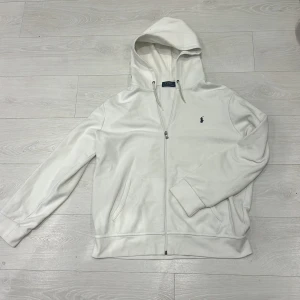 Vit zip hoodie från Polo Ralph Lauren - Vit hoodie från Polo Ralph Lauren med dragkedja och huva. Klassisk broderad logga på bröstet och två fickor framtill. Tillverkad i mjuk bomull, perfekt för en avslappnad stil. Små fläckar på framsidan som knappast syns