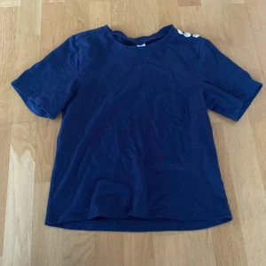 Mörkblå t-shirt med knappar på axeln från H&M Divided - Mörkblå t-shirt från H&M med tre guldiga knappar på ena axeln. T-shirten har rund halsringning och korta ärmar. Perfekt för en enkel och stilren look.