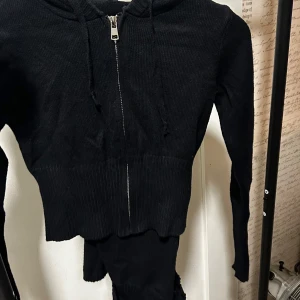 Svart croppad hoodie med dragkedja och tillkommande byxor - Säljer en svart croppad hoodie med dragkedja framtill och huva. Samt tillkommande byxor. Jackan har ribbad struktur och långa ärmar. Även byxorna har en ribbad struktur med silver detaljer vid midjan. Perfekt för en avslappnad och trendig stil.