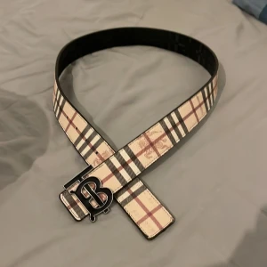 Vändbart bälte från Burberry med TB-spänne - Säljer ett vändbart bälte från Burberry med klassiskt rutigt mönster på ena sidan och svart krokopräglat skinn på andra. Stort metallspänne med TB-logga framtill. Perfekt accessoar för att lyfta din outfit.110cm