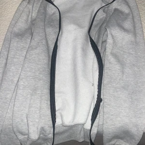 Ljusgrå ziptröja - Ljusgrå ziptröja med svarta detaljer längs dragkedjan. Mjuk och enkel design, perfekt för lager-på-lager. Långärmad och i ett bekvämt material.