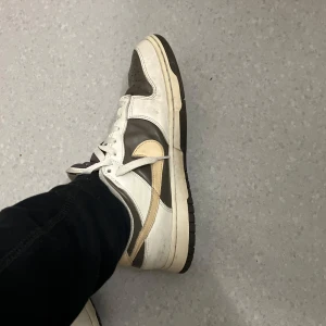 Nike Dunk Low i brunt och vitt - Säljer ett par Nike Dunk Low sneakers i brunt och vitt med beige swoosh. Skorna har klassisk snörning, rund tå och platt sula. Perfekta för dig som gillar streetwear och vill ha en stilren sneaker med retrovibbar.