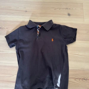 Pikétröja från Polo Ralph Lauren - Säljer en mörkblå pikétröja från Polo Ralph Lauren med klassisk krage och korta ärmar. Tröjan har en orange broderad logga på bröstet och knäppning vid halsen. Perfekt för en stilren och avslappnad look. Passar folk runt 150cm lång.