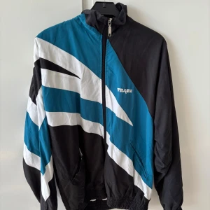 Svart och blå vindjacka från Track - Retro vindjacka från Track i svart, blått och vitt med grafiskt mönster. Jackan har dragkedja framtill, hög krage och resår vid ärmslut och midja. Tillverkad i polyester och nylon, perfekt för en sportig stil.