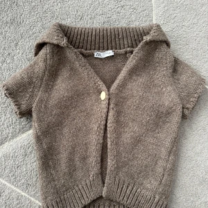 Brun stickad kofta från Zara - Kortärmad brun stickad kofta från Zara med en guldig knapp framtill och bred krage. Tröjan har ribbade muddar vid ärmslut och nederkant, perfekt för lager-på-lager-stil. Enkel och styla och har en fin touch. Pris kan diskuteras!