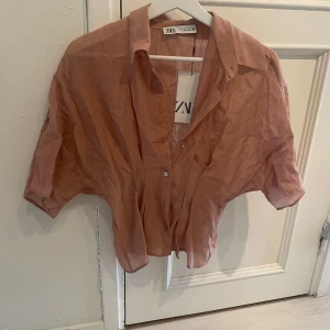 Vintagerosa croppad skjorta från Zara - Säljer en ljusrosa croppad skjorta från Zara med korta ärmar och klassisk krage. Skjortan är tunn och luftig, perfekt för varmare dagar. Stängs med knappar framtill och har en avslappnad passform.