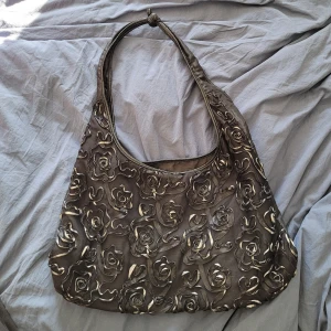 Svart axelväska med rosdetaljer - Säljer en svart axelväska med silvriga rosdetaljer i tyg på framsidan. Väskan har en mjuk form och dragkedja upptill. Tror märket är bulaggi, står det inne i väskan i alla fall.