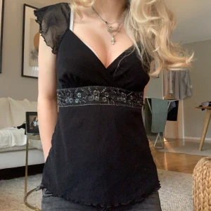 Svart omlottopp med spetsdetaljer - Svart omlottopp med transparenta volangärmar och v-ringning. Midjan har ett brett band med broderade blommor och knyt i ryggen. Skitt snygg o passar xs-s