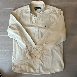 Beige Polo Ralph Lauren skjorta  - Klassisk beige skjorta från Polo Ralph Lauren med lång ärm och button-down krage. Skjortan har broderad logga på bröstet och knäppning framtill. Tillverkad i 100% bomull för en skön känsla.
