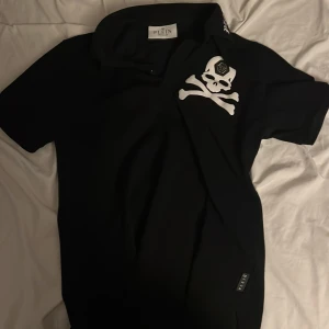 Svart philip plein piké  - Säljer en svart t-shirt från Plein med stor vit döskalle och korslagda ben på bröstet. Klassisk passform med korta ärmar och rund hals. Märkesdetalj på döskallens panna. Perfekt för dig som gillar edgy stil.