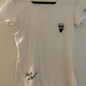 Vit t-shirt från Karl Lagerfeld med tryck - Vit t-shirt från Karl Lagerfeld med en liten tecknad patch framtill och broderad signatur på framsidan. Märkesnamnet är broderat på ärmen. Klassisk rund hals och korta ärmar. Perfekt för en stilren look.