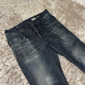 Mörkblå jeansbyxor från G-Star RAW - Säljer ett par mörkblå jeansbyxor från G-Star RAW med slitna detaljer och knappar i gylfen. Jeansen har klassisk femficksdesign och en brun läderpatch med logga bak i midjan. 