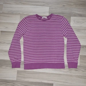 Lila randig tröja från H&M - Säljer en lila långärmad tröja från H&M med vita horisontella ränder. Tröjan har rund halsringning och ribbade muddar vid ärmslut och nederkant. Perfekt för dig som gillar färg och mönster!