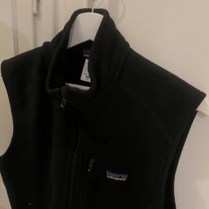 Svart fleeceväst från Patagonia - Svart fleeceväst från Patagonia med hög krage och dragkedja framtill. Västen har en bröstficka med dragkedja och två sidofickor. Perfekt för lager-på-lager och outdoor-vibes. På nära håll kan man se tecken på användning men fortfarande fräscht skick! 