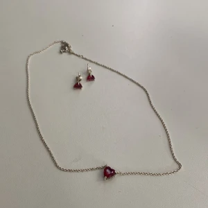 Pandora halsband & örhängen med hjärtan - Säljer ett sött set från Pandora med ett halsband och matchande örhängen i äkta silver. Båda har röda hjärtformade stenar som hänger i tunna kedjor. Perfekt för dig som gillar romantiska detaljer och vill ha något unikt och stilrent.