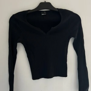 Svart ribbad topp från Gina Tricot - Säljer en svart, croppad långärmad topp från Gina Tricot i storlek XS. Toppen är ribbad med en snygg v-ringning framtill och har en tight passform. Perfekt att styla med höga jeans eller kjol för en trendig look.