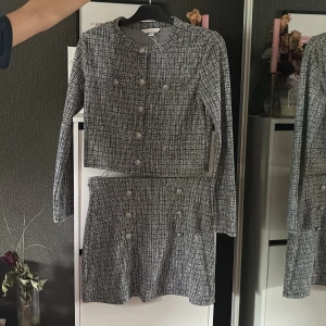 Grå kavaj och kjol set med knappar - Snyggt tvådelat set med kavaj och kjol i grått och vitt mönster. Kavajen har rund halsringning, långa ärmar och dekorativa knappar framtill. Kjolen är kort och har matchande knappar. Perfekt för en chic och trendig look.