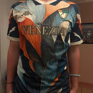 Venezia FC x Kappa fotbollströja - Unik Venezia FC fotbollströja från Kappa med korta ärmar. Tröjan har ett färgstarkt bladliknande mönster i blått, orange, beige och vitt. Stora tryck med klubbnamn och logga framtill. Tillverkad i 100% polyester, perfekt för dig som vill sticka ut på planen.