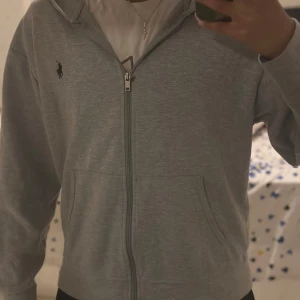 Grå hoodie från Polo Ralph Lauren - Grå hoodie från Polo Ralph Lauren med dragkedja framtill och klassisk svart logga på bröstet. Mjuk bomullskänsla, känguruficka och huva. Perfekt för chill dagar eller när du vill ha en clean och enkel stil.