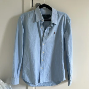 Ljusblå skjorta från Ralph Lauren S - Klassisk ljusblå skjorta från Ralph Lauren i custom fit. Skjortan har lång ärm, knappar framtill och ikonisk broderad logga på bröstet. Tillverkad i mjuk bomull och passar perfekt till en clean och stilren look.