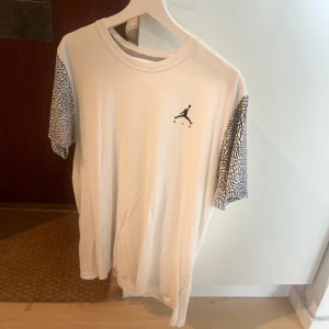 Vit Jordan t-shirt med mönstrade ärmar - Säljer en vit t-shirt från Jordan med det klassiska Jumpman-loggan på bröstet. Ärmarna har ett unikt svartvitt mönster som ger tröjan en sportig vibe. Perfekt för dig som gillar streetwear och vill sticka ut lite extra.