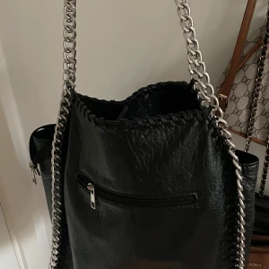Svart axelväska med kedja från Tiamo - Snygg svart axelväska från Tiamo i skinnimitation med grova silverfärgade kedjor som axelremmar. Väskan har en dragkedjeficka på framsidan och dekorativ söm längs kanten. Perfekt för dig som gillar en edgy och trendig look.