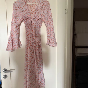 Adoore klänning rosa/lila  - Superfin långklänning med små blommor på rosa/lila botten, v-ringning och knyt framtill. Klänningen har långa volangärmar och ett lätt, flowigt tyg. Använd 2 h en midsommar. Aldrig tvättad. Fläckfri och nyskick. Kan mötas Ekerö annars skickar jag och då står köparen för frakten. 