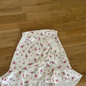 Blommig omlottkjol från NLY One - Supersöt vit omlottkjol från NLY One med rosa blommor och volangkant. Kjolen har en feminin och somrig vibe, perfekt för varma dagar. Knytband i midjan för extra detalj och snygg passform. Säljes i storlek XS. 