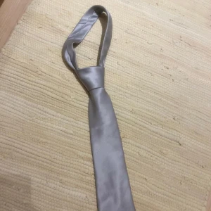 Silverfärgad slips från Dressmann - Snygg silverfärgad slips från Dressmann med diskret diagonal struktur. Slipsen har en klassisk form och är tillverkad i ett glansigt syntetmaterial som ger en stilren look. Perfekt för att lyfta din outfit med en elegant touch.