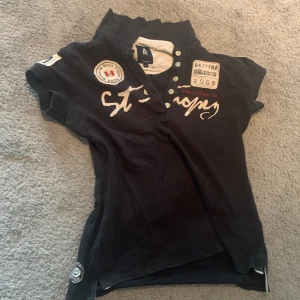 Svart kortärmad t-shirt från Gaastra - Snygg svart t-shirt från Gaastra med korta ärmar och coola broderade detaljer samt tryck på bröstet. Har knappar vid halsen och flera patchar med text och loggor. Perfekt för dig som gillar seglarstil och vill sticka ut.