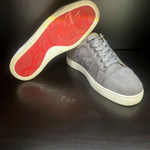 Grå mockasneakers från Louboutin - Snygga grå sneakers från Christian Louboutin i mjuk mocka med klassisk röd sula och vita kanter. Skorna har rund tå, snörning och diskreta metalldetaljer på sidan. Insidan är fodrad i beige läder för extra komfort. Perfekta för dig som gillar exklusiv streetstyle.