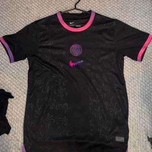 PSG x Nike svart fotbollströja - Snygg svart PSG fotbollströja från Nike med diskret blommönster i tyget. Tröjan har rosa och lila detaljer vid krage och ärmslut samt PSG-logga och Nike swoosh i rosa framtill. Perfekt för dig som vill sticka ut på planen eller på stan.
