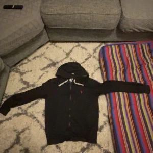 Svart hoodie från Ralph Lauren - Svart hoodie från Ralph Lauren med dragkedja och huva. Klassisk design med vit snörning i huvan och röd broderad logga på bröstet. Långärmad och tillverkad i mjuk bomull, perfekt för en avslappnad stil.