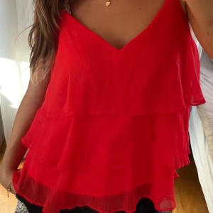 Röd volangig linnetopp - Säljer en röd linnetopp med flera lager volanger och smala axelband. Toppen har v-ringning och är tillverkad i ett lätt, luftigt material som ger en flowy känsla. Perfekt för dig som vill sticka ut med färg och volym. Lappen sitter kvar, aldrig använd!