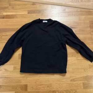Svart sweatshirt från Stone Island - Svart sweatshirt från Stone Island med broderad logga på bröstet. Klassisk rund halsringning och långa ärmar. Tillverkad i mjuk bomull som är skön att ha på sig. Perfekt för dig som gillar stilrena och enkla plagg med premiumkänsla.